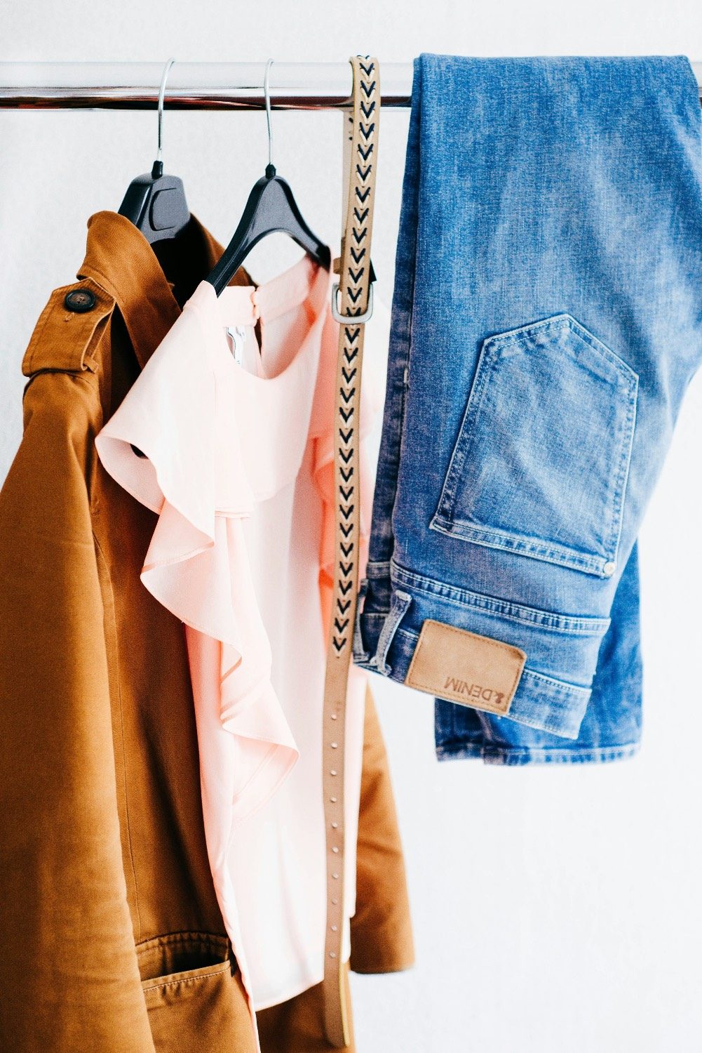 A tan jacket, a pink ruffled blouse, a braided belt and jeans on a clothes rail